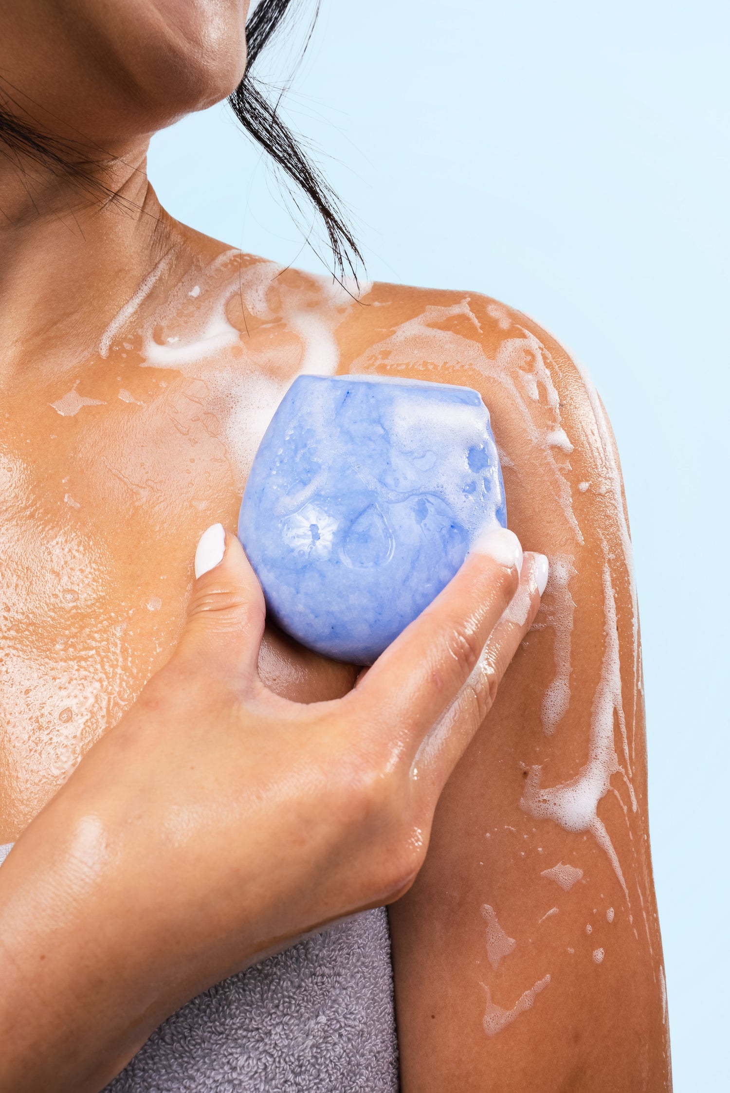 Female model holding a soapy blue body wash bar against her skin on a blue background.