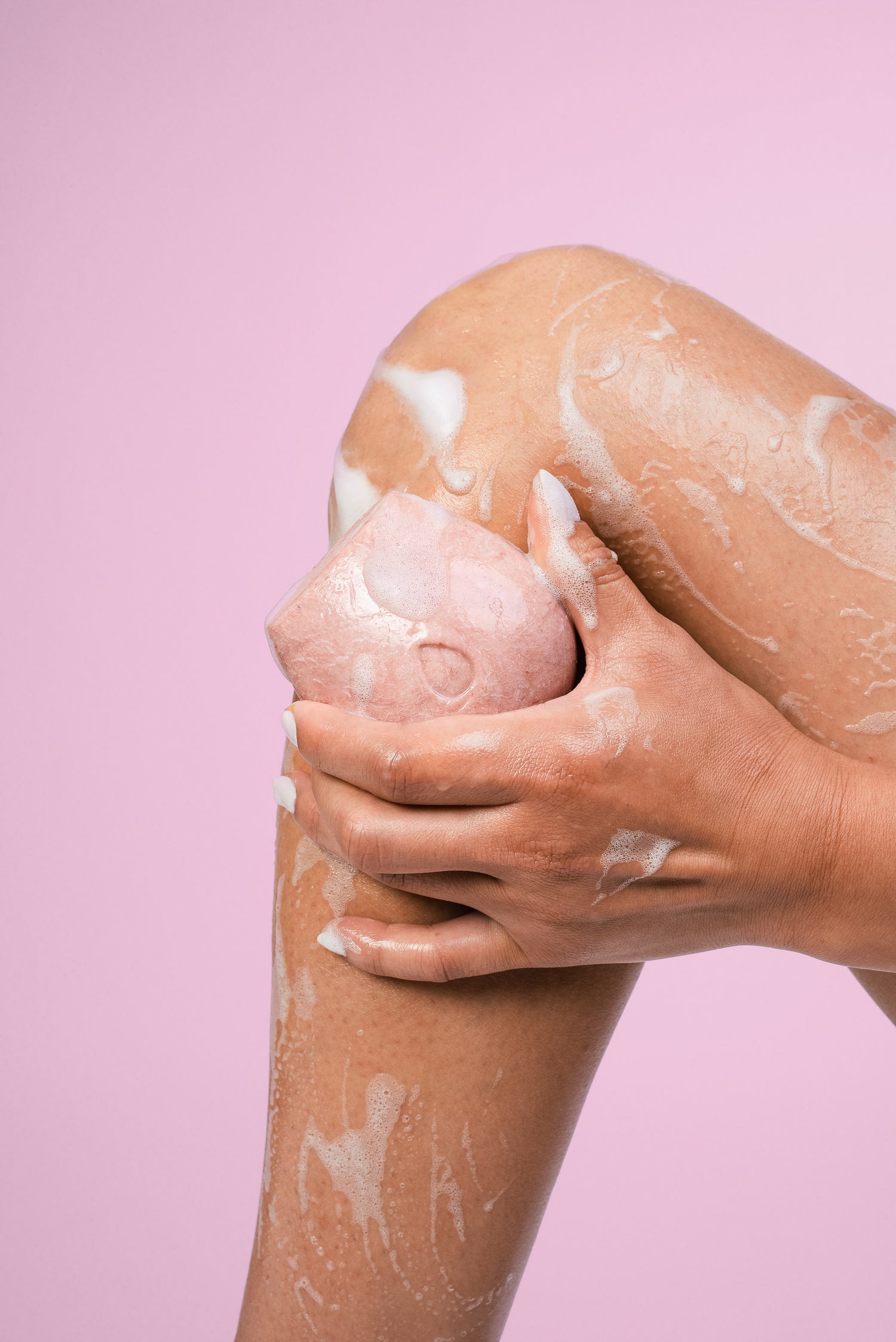 Model holding a wet pink body wash bar on her leg against a pink background.