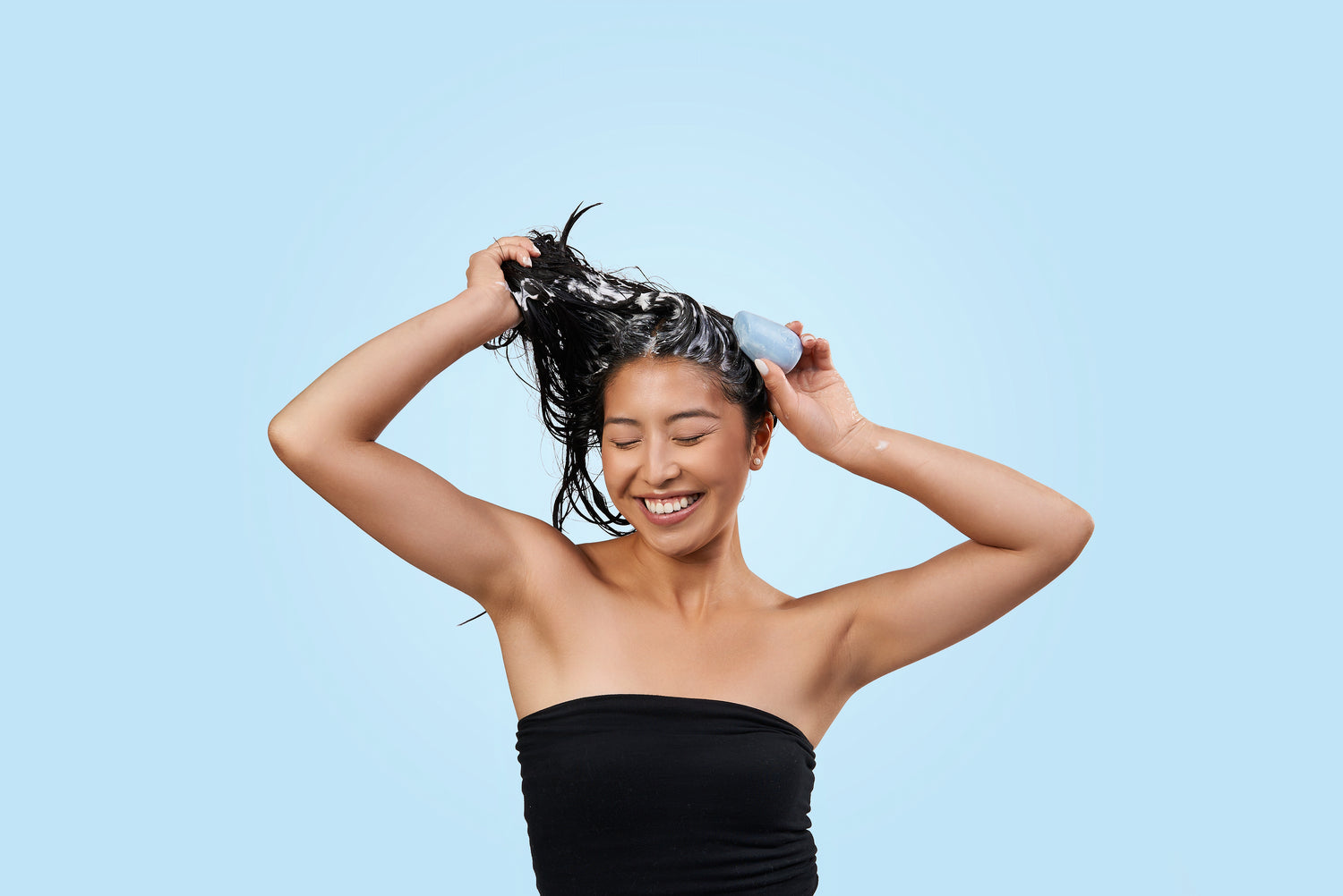 Female model with black hair using a blue shampoo bar on her hair against a blue background.