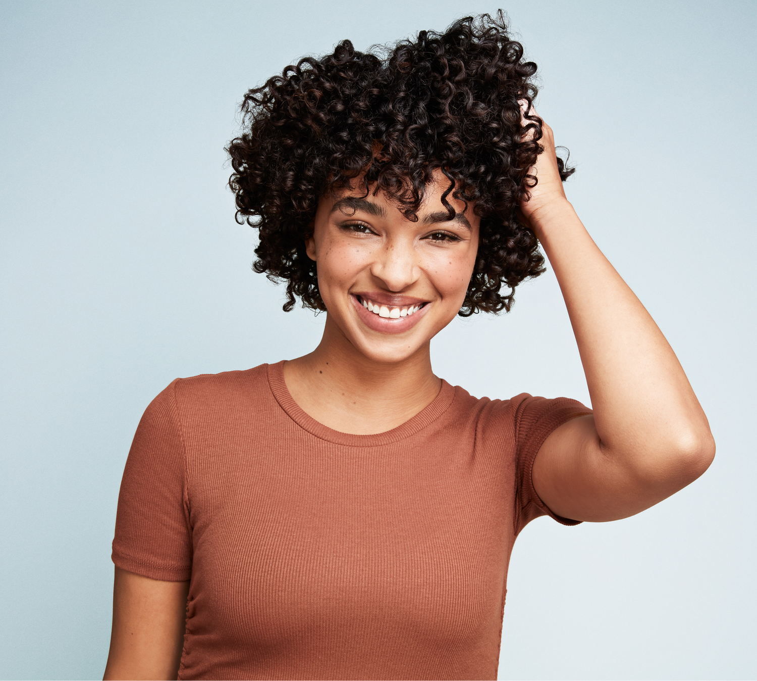 Woman with black curly hair smiling on a gray background.