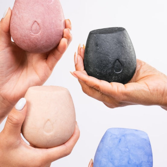 Two pairs of hands holding four different kinds of body wash bars on a white background.