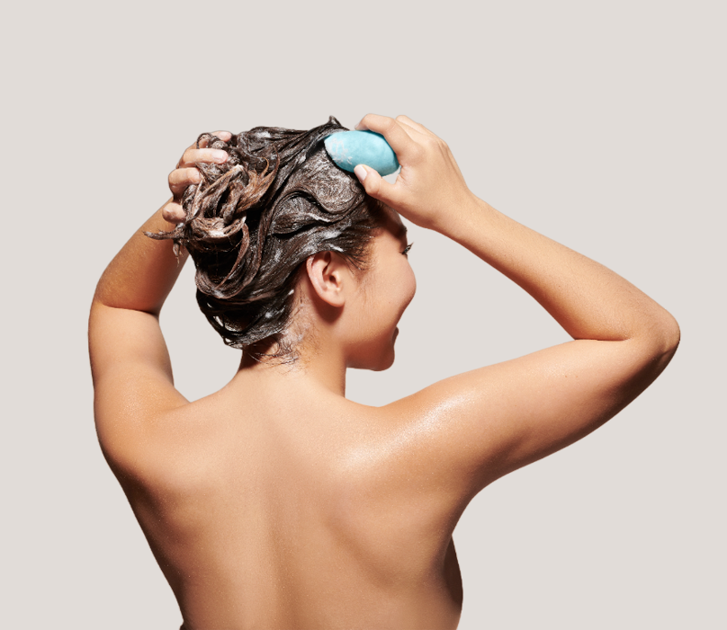 Woman with dark hair washing her hair with a blue shampoo bar on a white background.